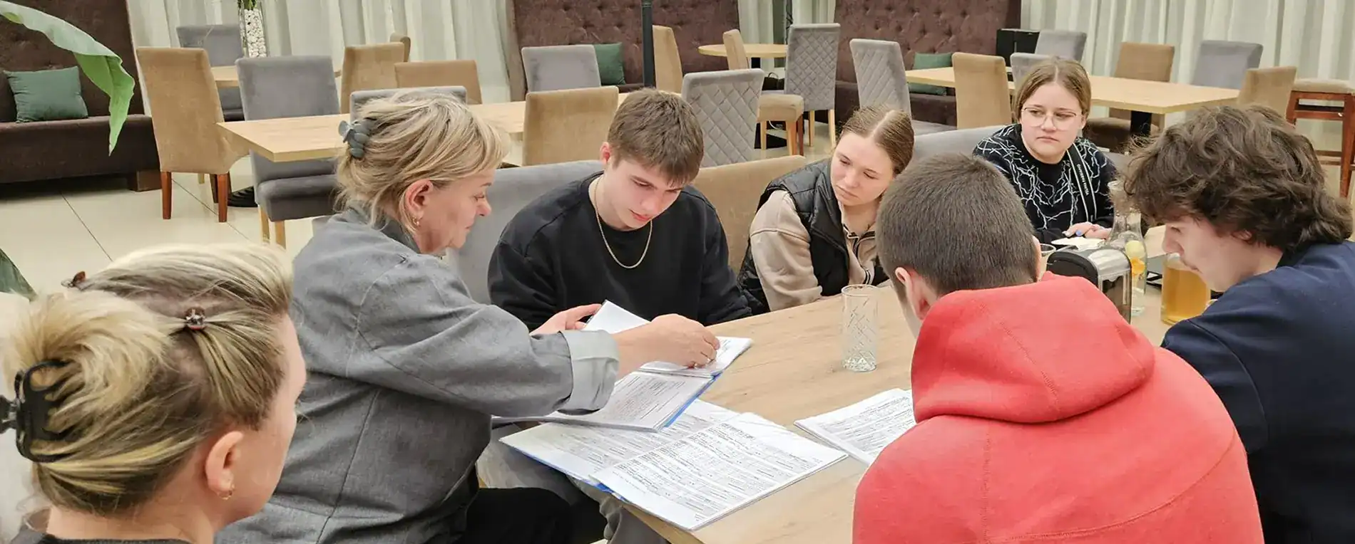 Šešių žmonių grupė sėdi prie stalo ir atidžiai žiūri į dokumentus, o viena moteris rodo į puslapį, siūlydama diskusiją ar susitikimą neformalioje, erdvioje patalpoje.