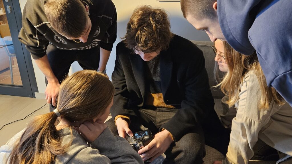 Penki jaunuoliai glaudžiai susibūrę aplink ant sofos sėdintį asmenį, kuris laiko fotoaparatą ir žiūri į jį. Visi jie atrodo smalsūs ir įsitraukę, įdėmiai stebi kamerą. Veiksmo vieta - patalpa.