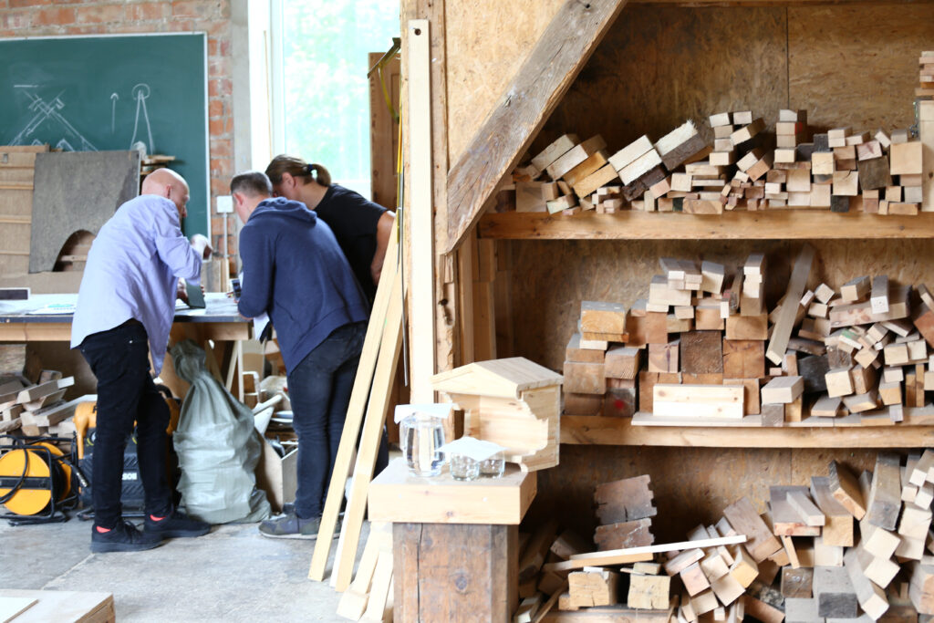 Medžio apdirbimo dirbtuvėje prie stalo dirba trys žmonės, apsupti medinių lentų krūvų, įrankių ir lentynų su įvairiomis medžio detalėmis.