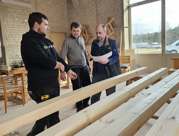 Trys vyrai stovi dirbtuvėse ir aptarinėja planus. Vienas jų laiko dokumentus, o du klausosi. Aplink juos matyti medinės sijos ir dirbtuvių įrankiai, o dideli langai praleidžia natūralią šviesą.